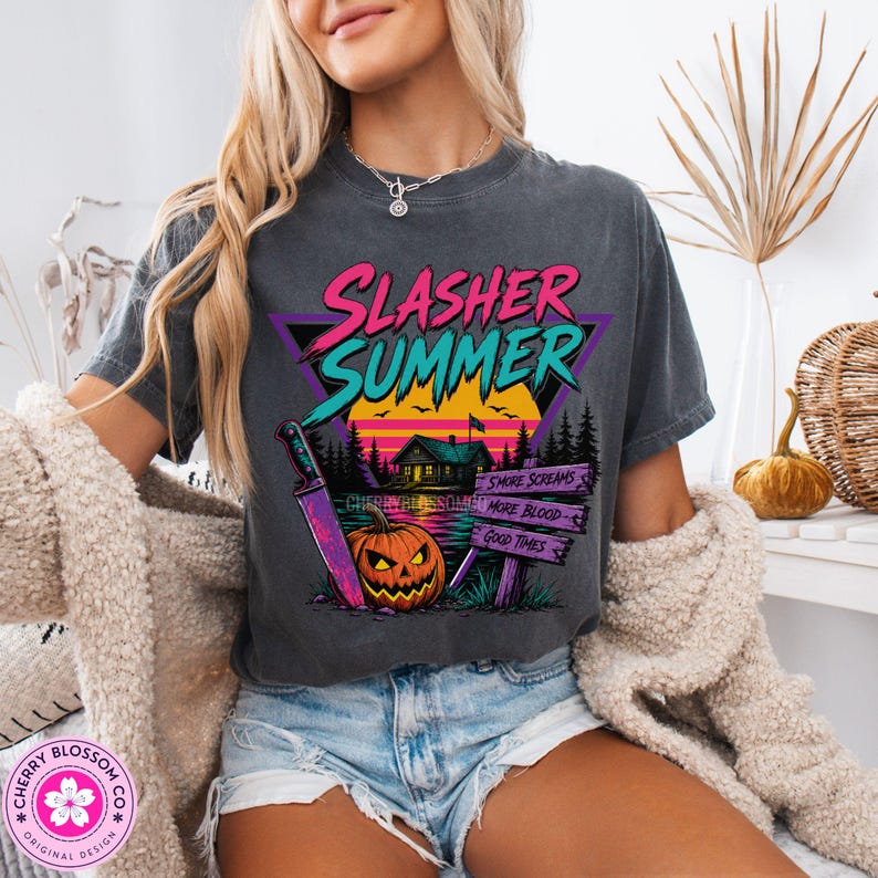 Puede incluir: Una camiseta gris carb&oacute;n de manga corta con un estampado gr&aacute;fico que incluye el texto Slasher Summer, una caba&ntilde;a, una calabaza, un cuchillo grande y un letrero con las palabras More Screams, More Blood y Good Times en colores vivos.