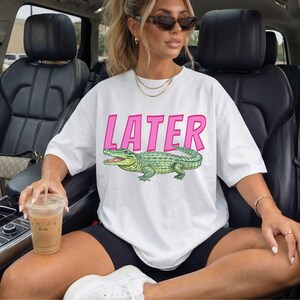 May include: White t-shirt featuring the word "LATER" in pink lettering and a green alligator graphic. A relaxed fit top, ideal for casual wear. The design is eye-catching.