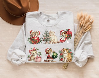 May include: A light gray sweatshirt featuring a festive Christmas design with six different illustrations of kittens. The kittens are depicted in various holiday scenes, including wearing Santa hats and decorating a Christmas tree.
