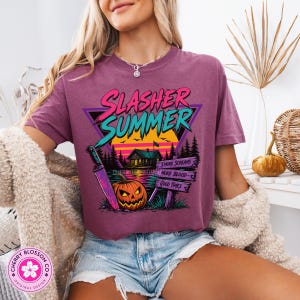 Puede incluir: Una camiseta color ciruela con un estampado gr&aacute;fico que incluye el texto "SLASHER SUMMER" en rosa y turquesa. El dise&ntilde;o muestra una caba&ntilde;a, una calabaza, un cuchillo grande y un letrero de madera con "SMORE SCREAMS", "MORE BLOOD" y "GOOD TIMES".