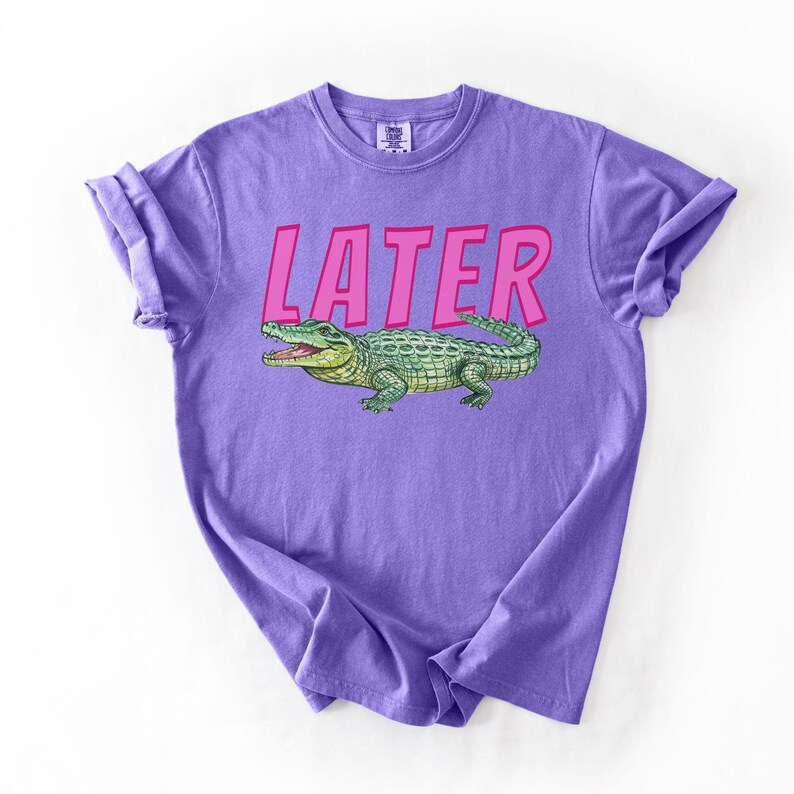 May include: A lilac t-shirt featuring the word "LATER" in pink lettering above a green and yellow alligator illustration. The t-shirt is folded with rolled-up sleeves.