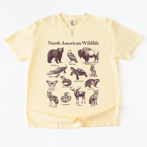 Pu&ograve; includere: T-shirt giallo pallido con una grafica della fauna selvatica nordamericana. Il design include illustrazioni di orso nero, aquila calva, bisonte, alligatore, tartaruga, gufo, farfalla, lontra di fiume, procione, coyote, serpente, alce e volpe rossa.