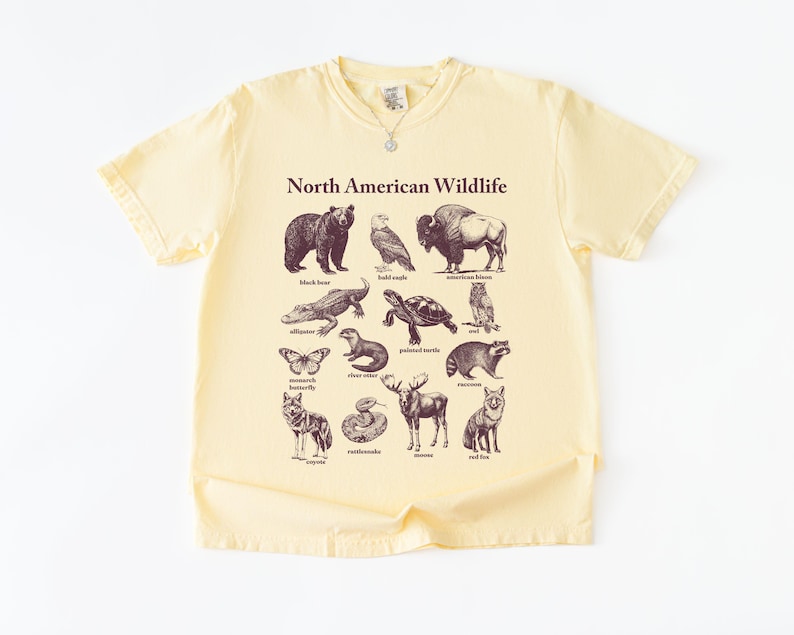 Pu&ograve; includere: T-shirt giallo pallido con una grafica della fauna selvatica nordamericana. Il design include illustrazioni di orso nero, aquila calva, bisonte, alligatore, tartaruga, gufo, farfalla, lontra di fiume, procione, coyote, serpente, alce e volpe rossa.