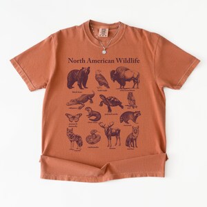 Pu&ograve; includere: T-shirt color ruggine con una grafica della fauna selvatica nordamericana. Il design include illustrazioni di un orso, un gufo, un bisonte, un alligatore, una tartaruga e altri animali. Le parole "North American Wildlife" sono stampate in alto.