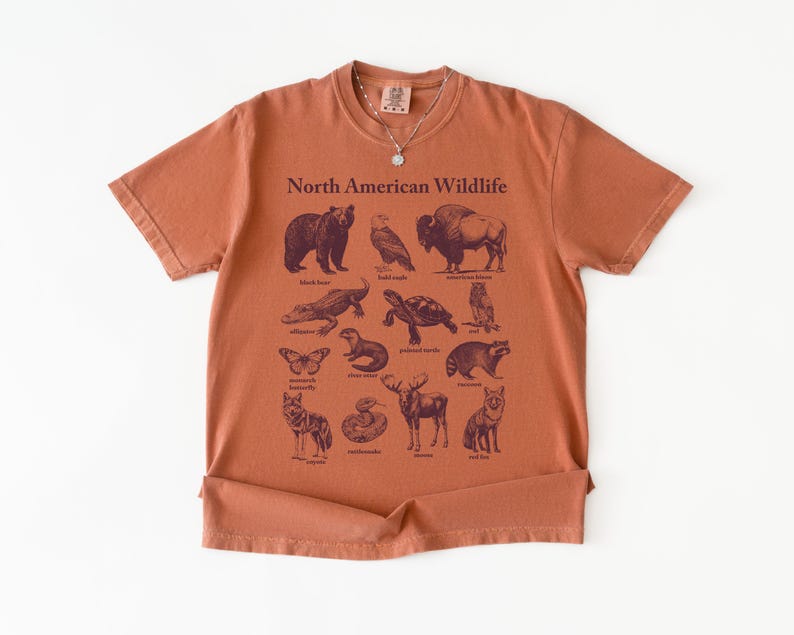 Pu&ograve; includere: T-shirt color ruggine con una grafica della fauna selvatica nordamericana. Il design include illustrazioni di un orso, un gufo, un bisonte, un alligatore, una tartaruga e altri animali. Le parole "North American Wildlife" sono stampate in alto.