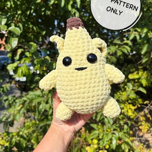 May include: A light yellow crochet plush toy shaped like a banana with a brown stem, two large black plastic eyes, and a small black stitched mouth. A white circular graphic in the upper right corner contains the text PDF PATTERN ONLY.