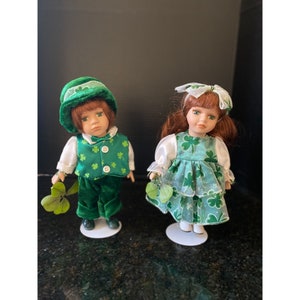 May include: Two porcelain dolls dressed in green shamrock print outfits. The boy doll wears a green hat and holds a shamrock. The girl doll wears a green dress with a white bow and holds a shamrock.