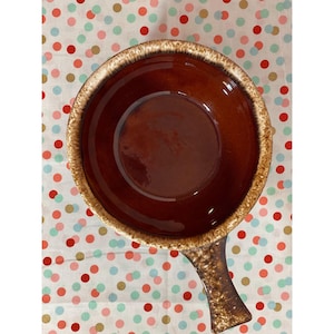 May include: A brown ceramic soup bowl with a handle. The bowl has a glossy interior and a textured, light brown rim. The handle is also brown with a textured design. The bowl is set against a polka dot background.