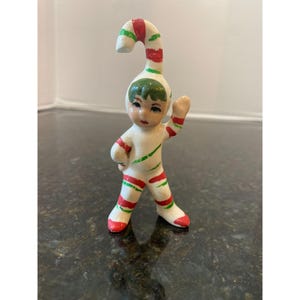 Puede incluir: Pequeña figura de cerámica de un niño vestido de bastón de caramelo. La figura es blanca con rayas y detalles rojos y verdes. El niño tiene una gorra verde y saluda con una mano. La figura es una decoración festiva navideña.
