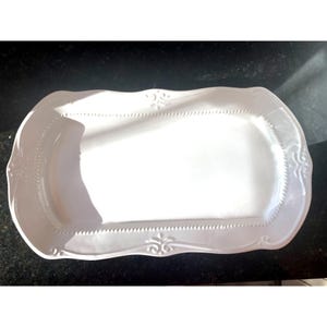 May include: A white, rectangular serving platter with a scalloped edge and ornate detailing. The platter has a raised border with a dotted pattern and decorative flourishes at the corners. The surface is smooth and glossy.
