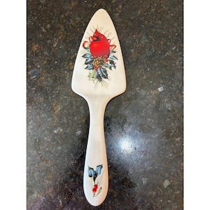 Può includere: Un server per torte in ceramica con manico e lama color crema. La lama presenta un disegno di uccello cardinale rosso con un nastro rosso, una pigna e un agrifoglio. Il manico ha un disegno floreale blu e rosso.