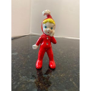 May include: A small, vintage ceramic figurine of a child in a red onesie and matching cap with a white pom-pom. The figurine has blonde hair, blue eyes, and is touching its cheek. The figure is standing on a dark surface.