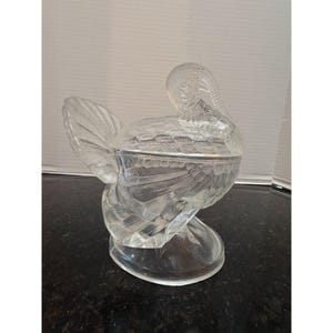 May include: A clear glass turkey-shaped decorative container with a lid. The container has detailed feather and body patterns. The turkey is standing on a clear, oval-shaped base. The lid is shaped like the turkey's head and neck.