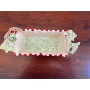 May include: A white ceramic serving dish with a red and white striped border. The dish is decorated with a green swirl pattern and features a 3D reindeer on one end and a 3D Christmas tree on the other. The tree has the word "Peace" on it.