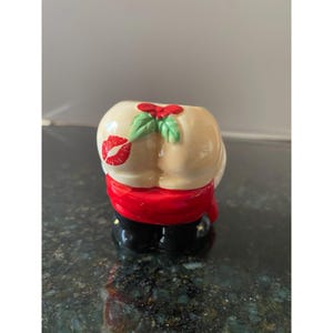 May include: A ceramic figurine of Santa Claus's backside with a red bow and a green holly sprig. The figurine is painted with a white background and a red kiss mark.