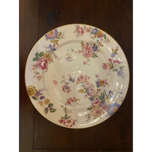 May include: A round, cream-colored plate with a gold rim. The plate is decorated with a floral pattern featuring pink, blue, and yellow flowers and green leaves. The plate is set on a dark brown wooden surface.