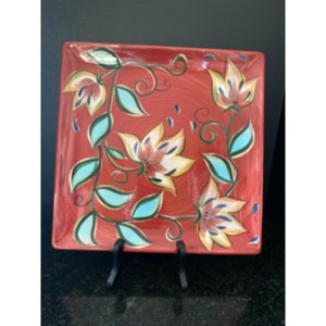 Puede incluir: Plato cuadrado de cerámica con fondo rojo y diseño floral. Las flores están pintadas en amarillo, naranja y blanco, con hojas y enredaderas turquesas. El plato está sostenido por un soporte de metal negro.