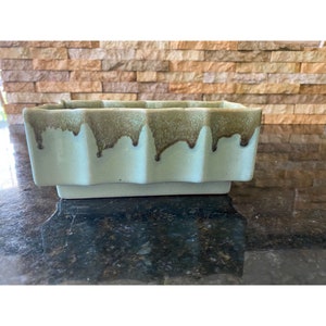 以下が含まれることがあります： 波打つ縁取りと、側面に垂れ下がった茶色の釉薬が施された、薄い緑色のセラミック製の植木鉢です。