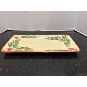 May include: Rectangular serving platter with a cream-colored center and a red border. The platter is decorated with green sprigs and red berries at each corner, creating a festive holiday design. The dish is made of ceramic.