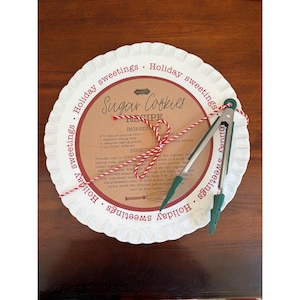 May include: A white scalloped plate featuring the text Holiday sweetings and a central brown label with a Sugar Cookies recipe. A pair of silver metal kitchen tongs with green silicone tips is tied to the plate with red and white twine.