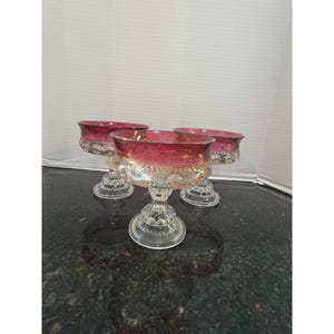 Puede incluir: Tres cuencos de postre de cristal transparente con borde rojo rubí. Los cuencos tienen una base de pedestal con un diseño decorativo. Los cuencos están sobre una superficie oscura.