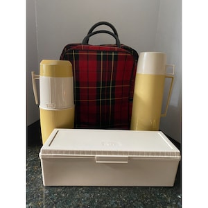 Puede incluir: Un juego de picnic vintage con una bolsa aislada de cuadros rojos y negros, dos termos blancos y amarillos y un contenedor de almacenamiento rectangular blanco.