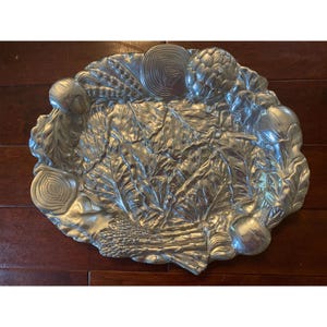 May include: A silver-colored, oval-shaped serving platter with a detailed vegetable design. The platter features embossed images of artichokes, onions, asparagus, and leafy greens. The platter is made of metal and is suitable for serving food.