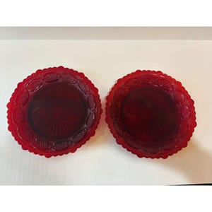 May include: Two ruby red glass plates with a scalloped edge and a detailed embossed pattern. The plates have a central design with vertical lines radiating from the centre. The plates are likely for serving or display.