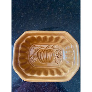 Könnte beinhalten: Eine braune Keramikform zum Backen eines Kürbis-Kuchen. Die Form hat ein erhabenes Design eines Kürbisses mit Blättern.
