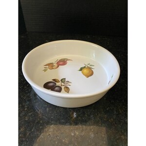 May include: White ceramic baking dish with a fruit design. The design features a red apple, a yellow lemon, and two purple plums.