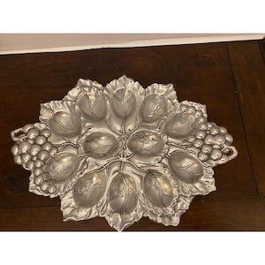 Op de afbeelding: Zilverkleurige serveerschaal met een druiven- en bladerontwerp. De schaal heeft een centraal gedeelte omgeven door bladvormige compartimenten en trossen druiven aan de zijkanten. De schaal is gemaakt van metaal en is ontworpen voor het serveren van voedsel.