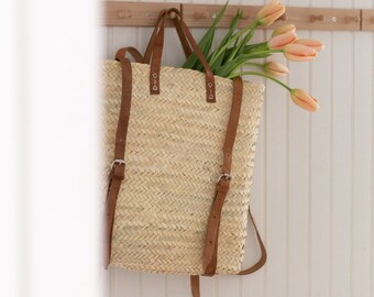 May include: A woven straw tote bag with brown leather straps and silver-toned buckles, featuring a rectangular shape and an open top. The bag is filled with a bouquet of light peach-colored tulips with green stems and leaves, hanging from a hook.
