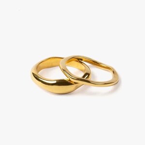 May include: Two gold rings. One is a thick band with a smooth, rounded shape. The second ring has a wavy, open design. Both rings are made of a shiny, reflective metal and are set against a white background.