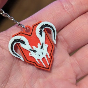 Puede incluir: Un llavero rojo y negro con un emblema blanco y negro. El emblema presenta un dise&ntilde;o estilizado con formas curvas y un contorno en forma de escudo. El llavero est&aacute; sujeto a una cadena plateada.