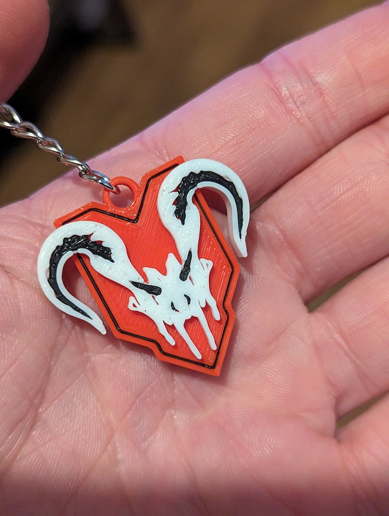 Puede incluir: Un llavero rojo y negro con un emblema blanco y negro. El emblema presenta un dise&ntilde;o estilizado con formas curvas y un contorno en forma de escudo. El llavero est&aacute; sujeto a una cadena plateada.
