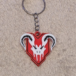 Puede incluir: Un llavero rojo y blanco con un dise&ntilde;o de calavera estilizada. El llavero tiene un anillo y una cadena de metal plateado. El dise&ntilde;o de la calavera est&aacute; sobre un escudo rojo. El llavero es probablemente de pl&aacute;stico.