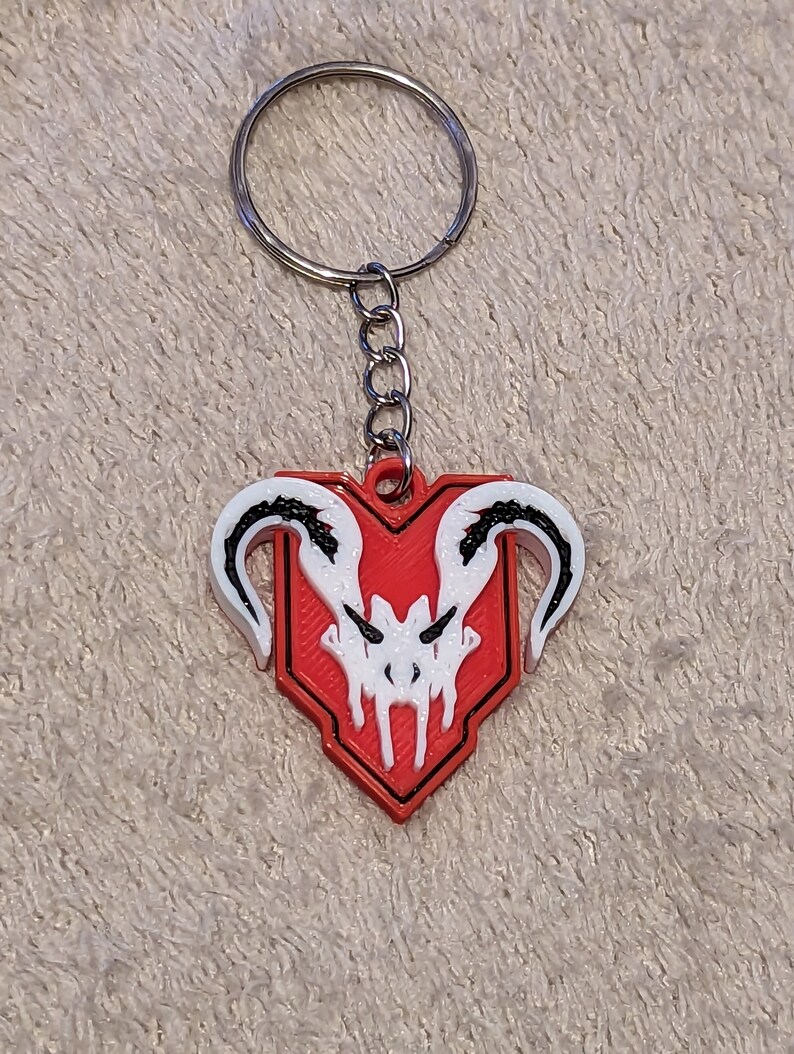 Puede incluir: Un llavero rojo y blanco con un dise&ntilde;o de calavera estilizada. El llavero tiene un anillo y una cadena de metal plateado. El dise&ntilde;o de la calavera est&aacute; sobre un escudo rojo. El llavero es probablemente de pl&aacute;stico.