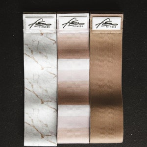 May include: Three fabric resistance bands with different patterns. The bands are white with a marble pattern, white with pink stripes, and brown. Each band has the text "Fitness" printed on a white label.