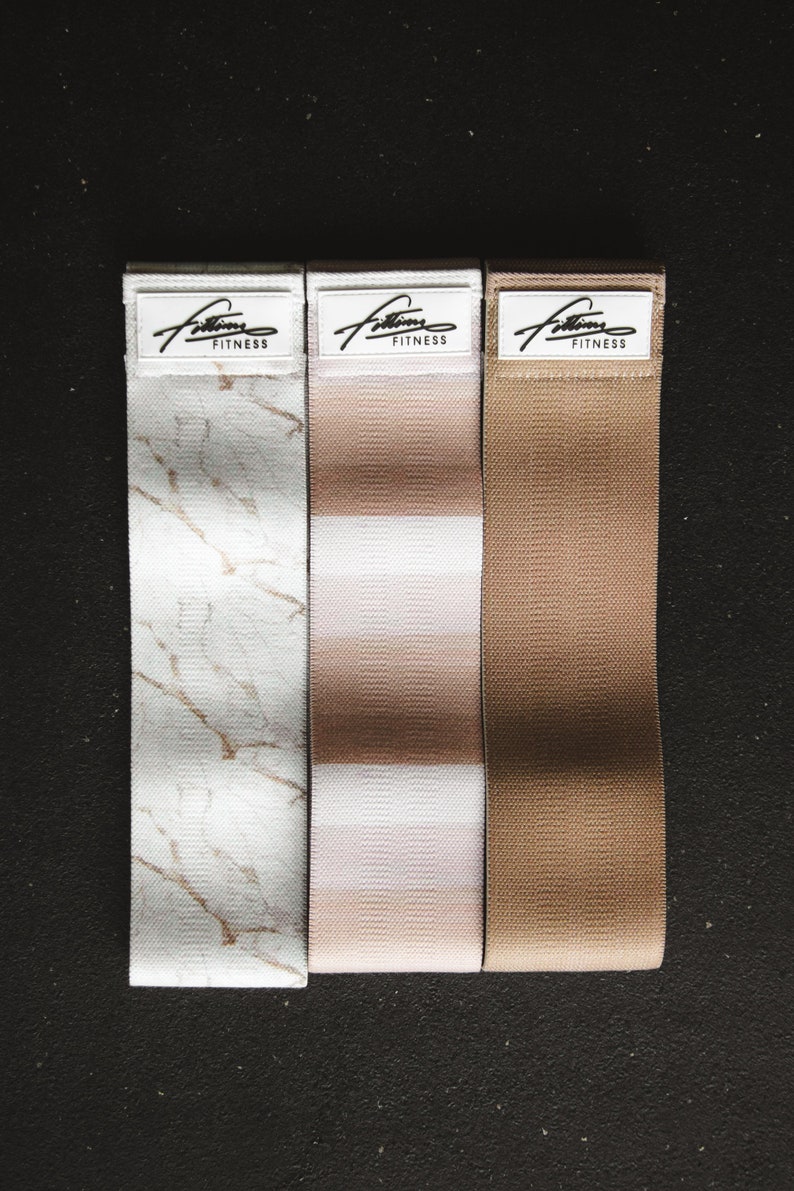 May include: Three fabric resistance bands with different patterns. The bands are white with a marble pattern, white with pink stripes, and brown. Each band has the text "Fitness" printed on a white label.