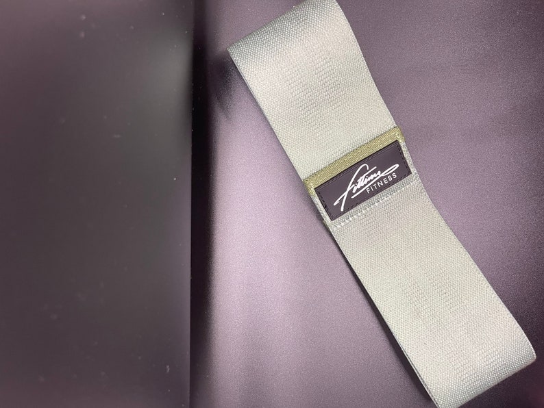 May include: A light gray fabric resistance band with a black label that says "Fitline Fitness".