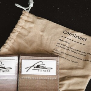 May include: A beige drawstring bag with the word "Consistent" printed on it. The bag is open and you can see two beige resistance bands with the word "Fitness" printed on them.