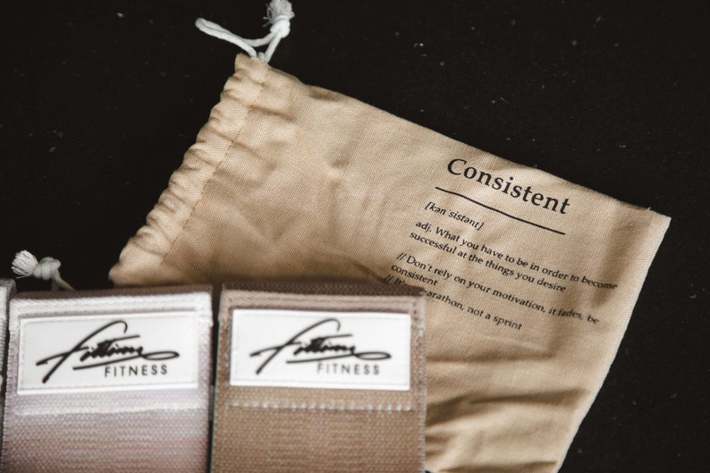 May include: A beige drawstring bag with the word "Consistent" printed on it. The bag is open and you can see two beige resistance bands with the word "Fitness" printed on them.