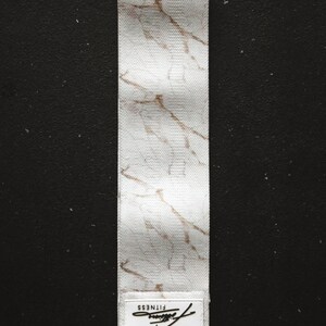 May include: White fabric resistance band with a marble pattern and gold accents. The band has a white label with the word "FITNESS" printed on it.