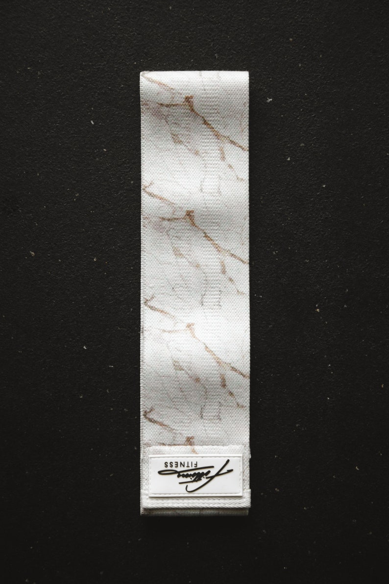 May include: White fabric resistance band with a marble pattern and gold accents. The band has a white label with the word "FITNESS" printed on it.
