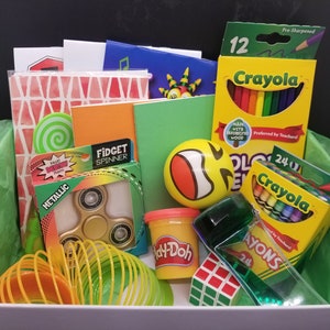 Op de afbeelding: Een witte doos gevuld met verschillende schoolbenodigdheden, waaronder kleurpotloden, een fidget spinner, klei, een slinky en een groene stressbal met een smiley. De doos is bekleed met groen tissuepapier.