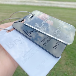 May include: Two luggage tags with black and white text. The top tag is clear and has the name "Briana" with the text "360 West 200 St Orlando FL 32804" and a phone number. The bottom tag is white and has the name "Brya" with the text "360 West 200 St Orlando FL 32804" and a phone number.