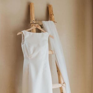 May include: A white wedding dress hangs on a wooden ladder with a white veil draped over the top rung. The dress has a simple, elegant design and is made of a smooth fabric. The hanger is personalized with the word "Bride".