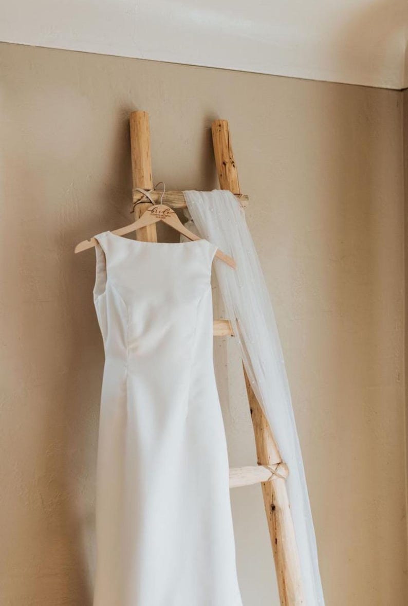 May include: A white wedding dress hangs on a wooden ladder with a white veil draped over the top rung. The dress has a simple, elegant design and is made of a smooth fabric. The hanger is personalized with the word "Bride".