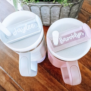 May include: Two personalized stainless steel travel mugs with lids and handles. The mug on the left is light blue with the name "Jaylee" on the lid. The mug on the right is pink with the name "Brooklyn" on the lid.