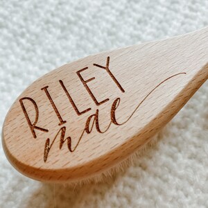 May include: A wooden hair brush with a natural bristle brush head. The handle is engraved with the name "Riley Mae" in a cursive font.